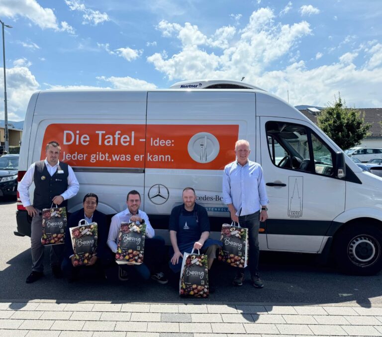 Gruppenfoto von fünf Menschen vor einem Lieferwagen der Tafel, die Einkaufstaschen von Edeka als Teil einer Spende halten.