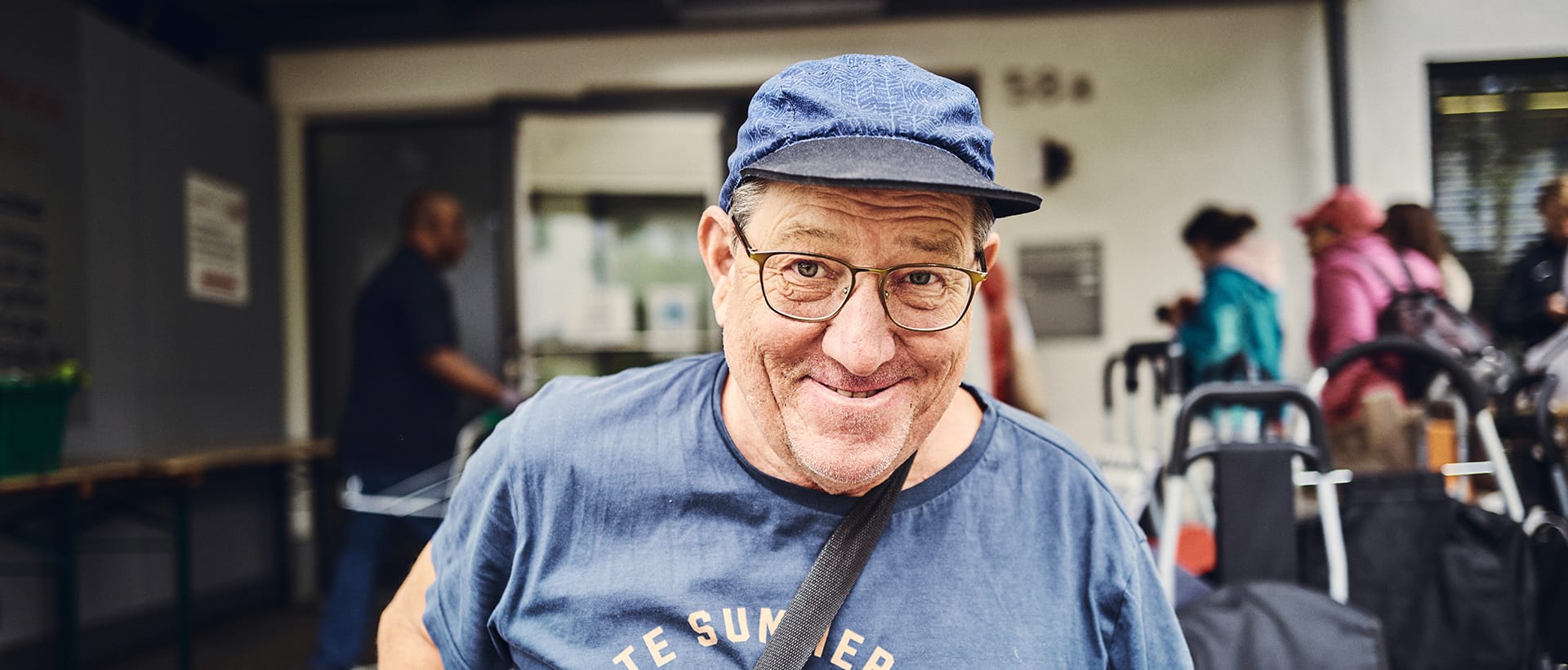 Ein lächelnder Mann mit Brille und Mütze steht vor dem Eingang.