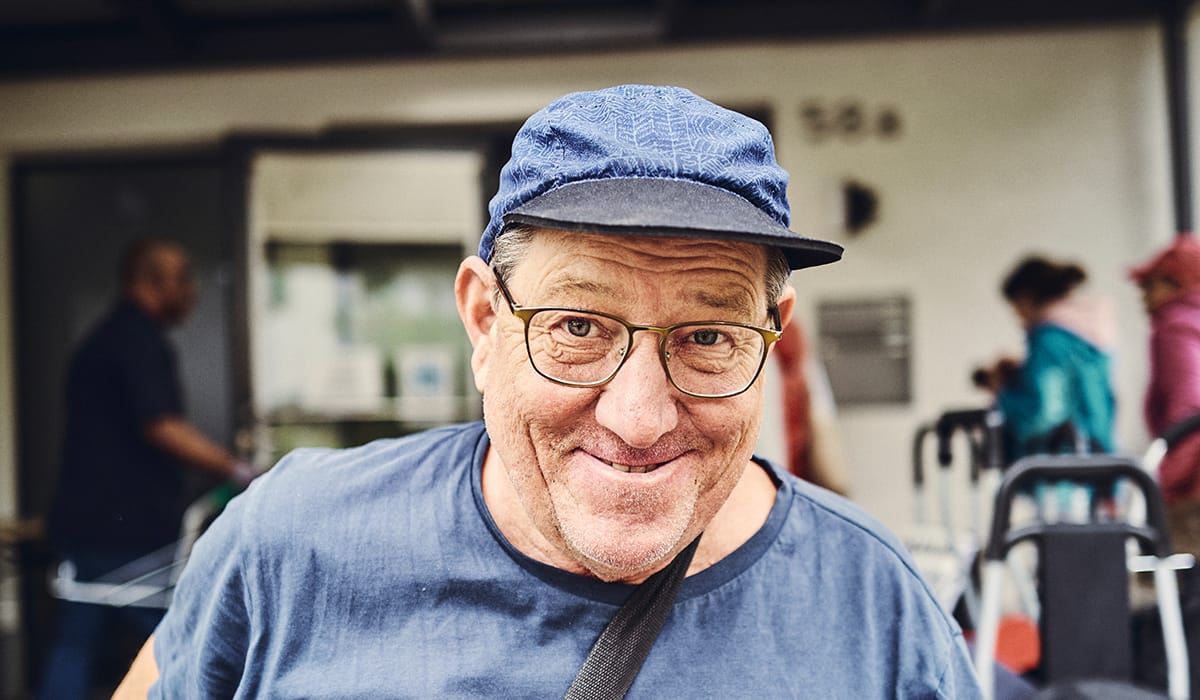 Ein lächelnder Mann mit Brille und Mütze steht vor dem Eingang.