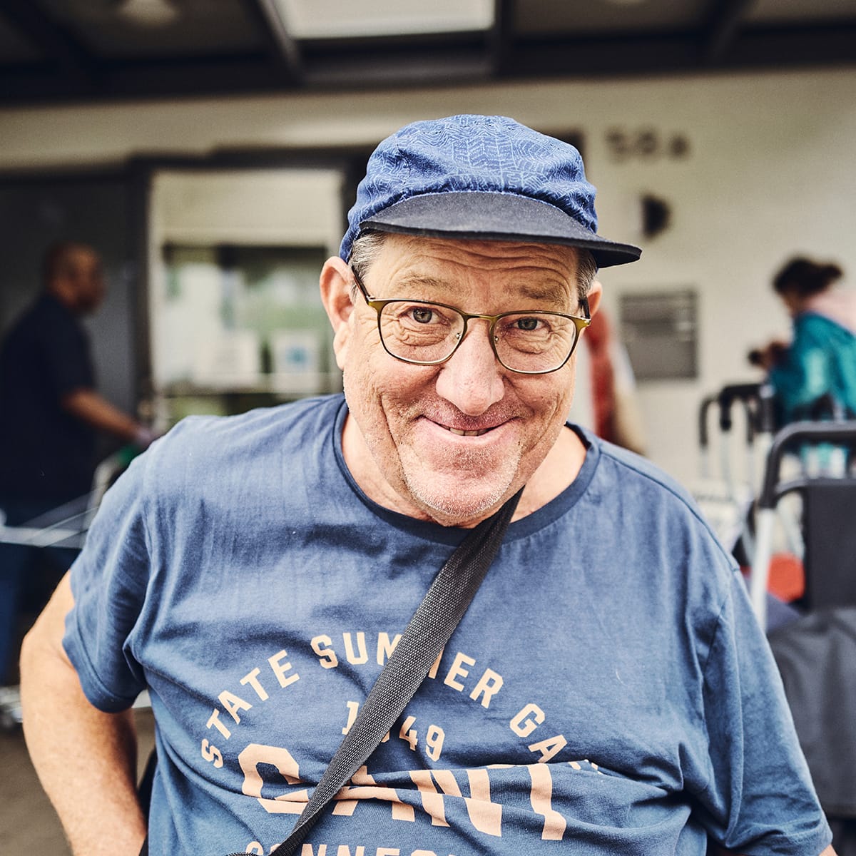 Ein lächelnder Mann mit Brille und Mütze steht vor dem Eingang.