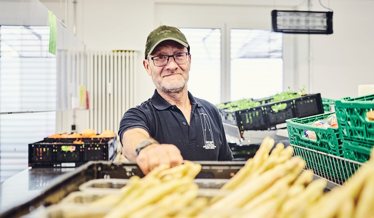 Ein Mann in einem Tafel-Shirt steht an einer Kiste mit frischem Spargel.