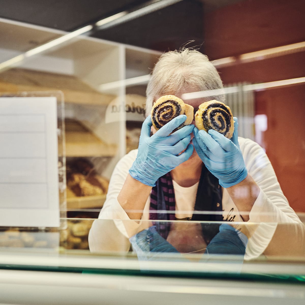 Eine Frau hält sich zwei Mohnschnecken humorvoll vor ihr Gesicht und sorgt für einen fröhlichen Moment hinter der Theke.